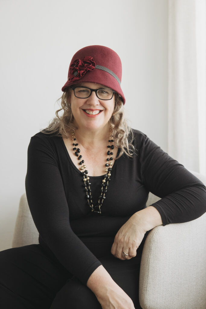 Author Ava Ness wearing a burgundy cloche hat with a flower detail and black-rimmed glasses, smiling slightly while sitting in a cream coloured arm chair. She has curly ash blonde hair and is dressed in a black top with a long beaded necklace.